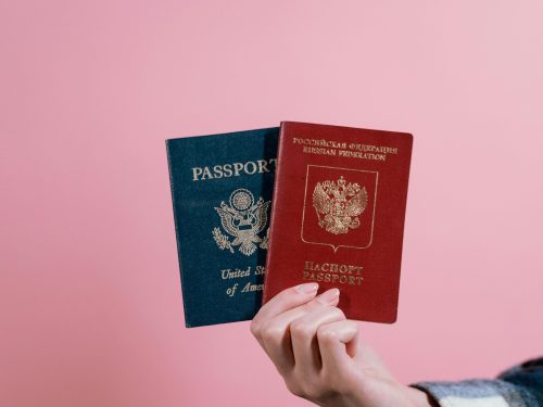 Close-up of a hand holding American and Russian passports on a pink background.